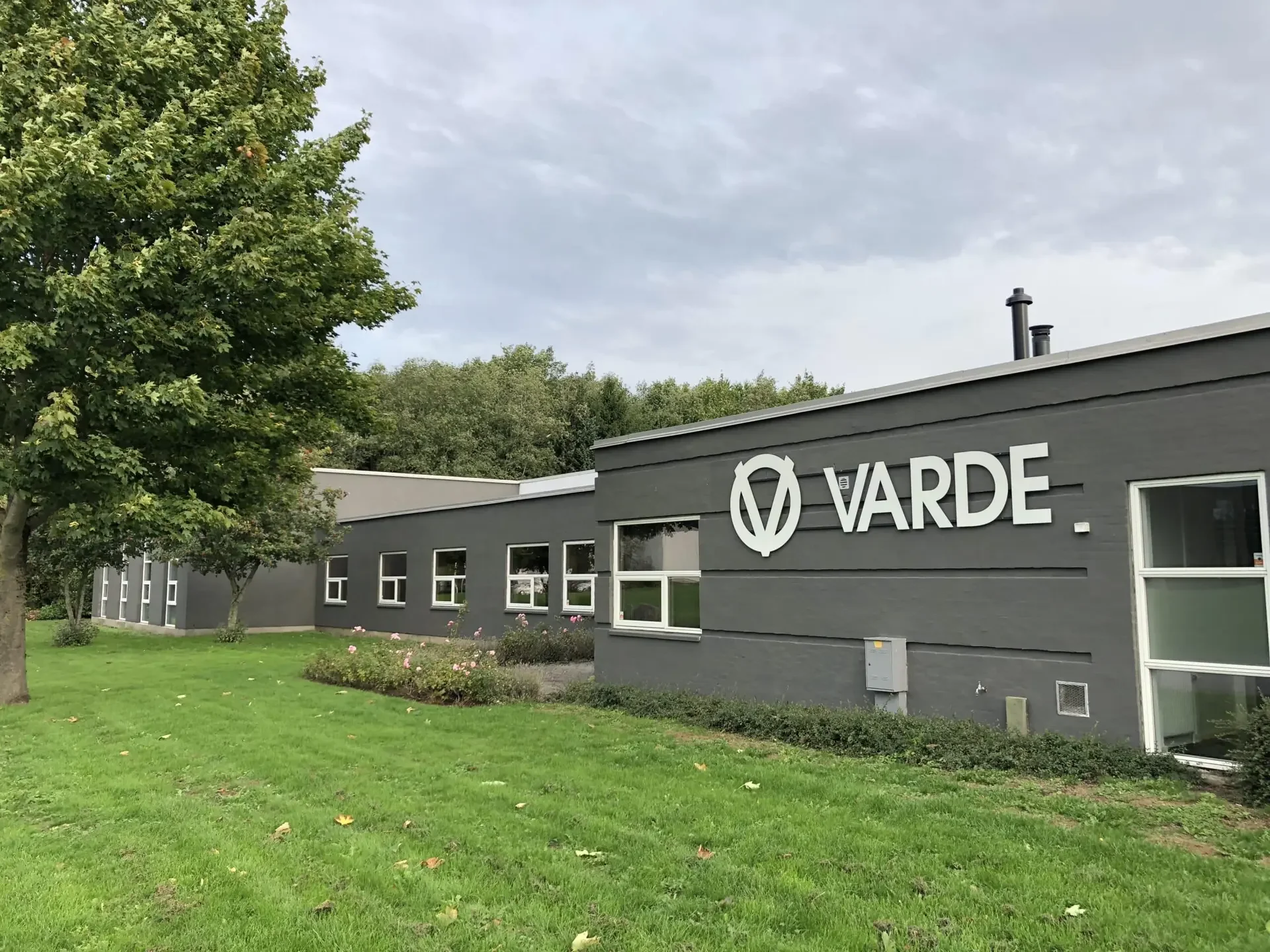 VARDE Fabrikgebäude mit Firmenschild, Rasen und Bäumen